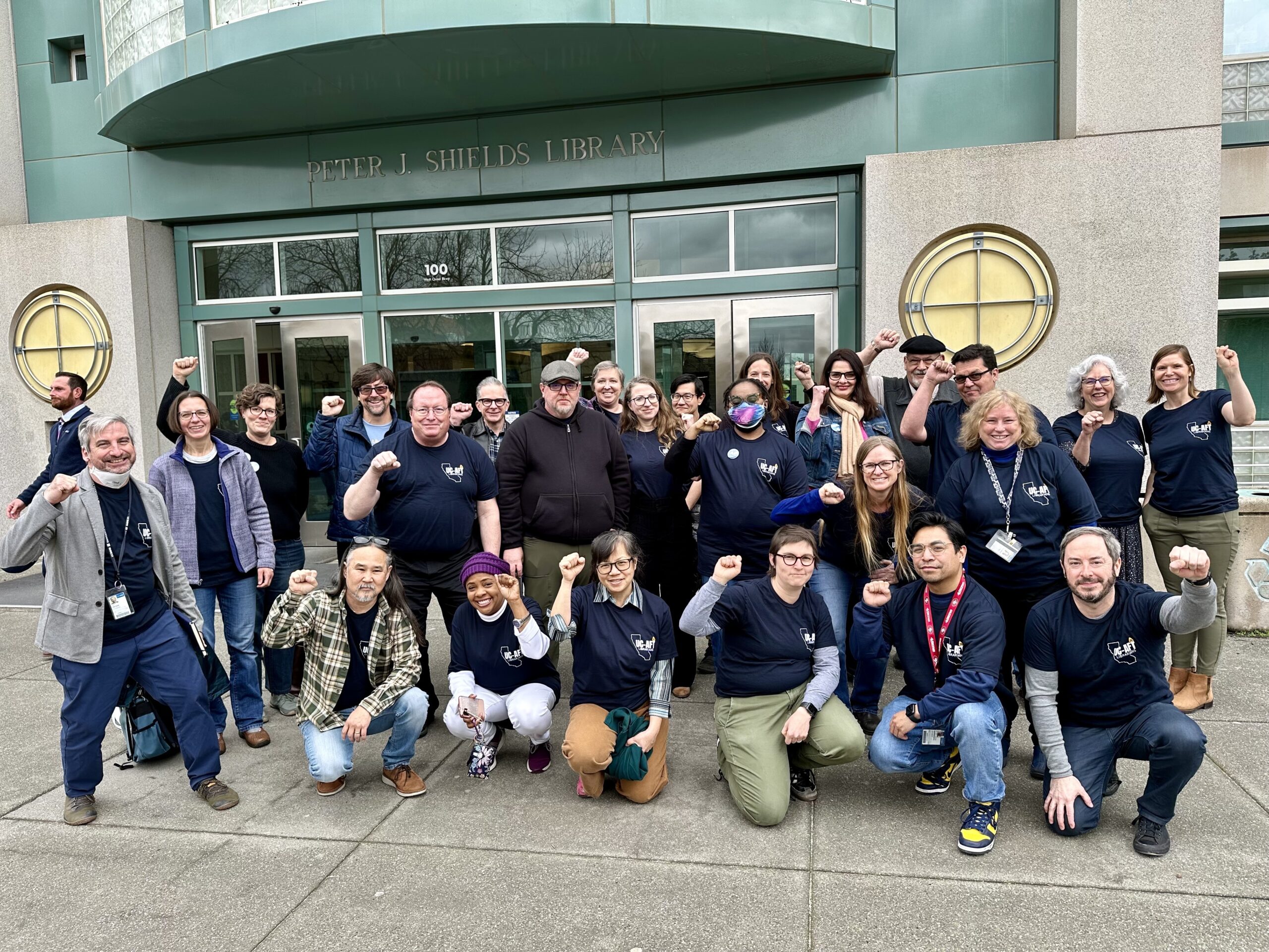 Group of librarians and allies in blue union t-shirts raise their fists in solidarity outside the Shields Library at UC Davis.