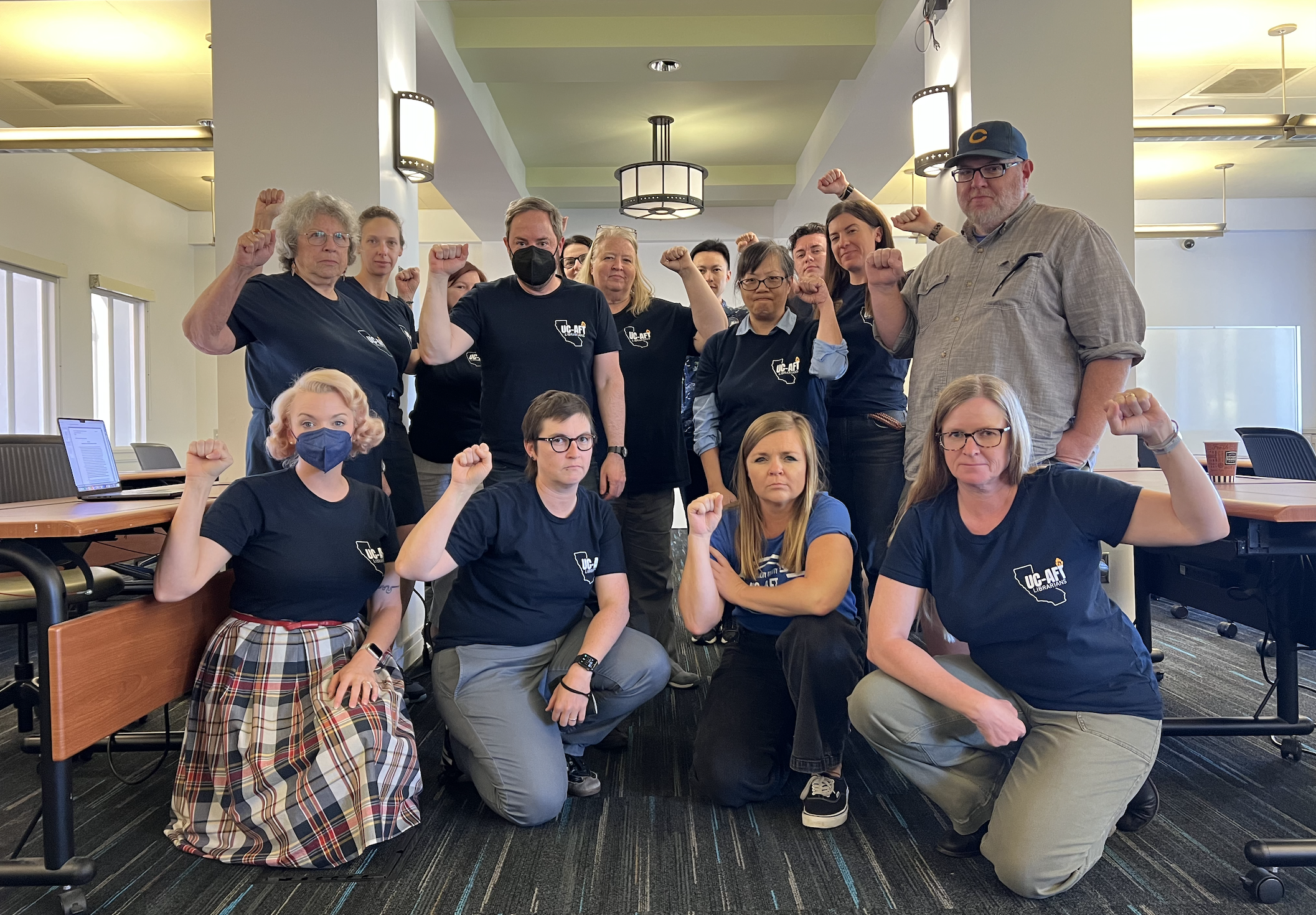 A group of people, both standing and kneeling, are posing indoors with serious expressions. They are raising their fists in a gesture of solidarity. The individuals are wearing matching dark blue T-shirts with a logo on the front.