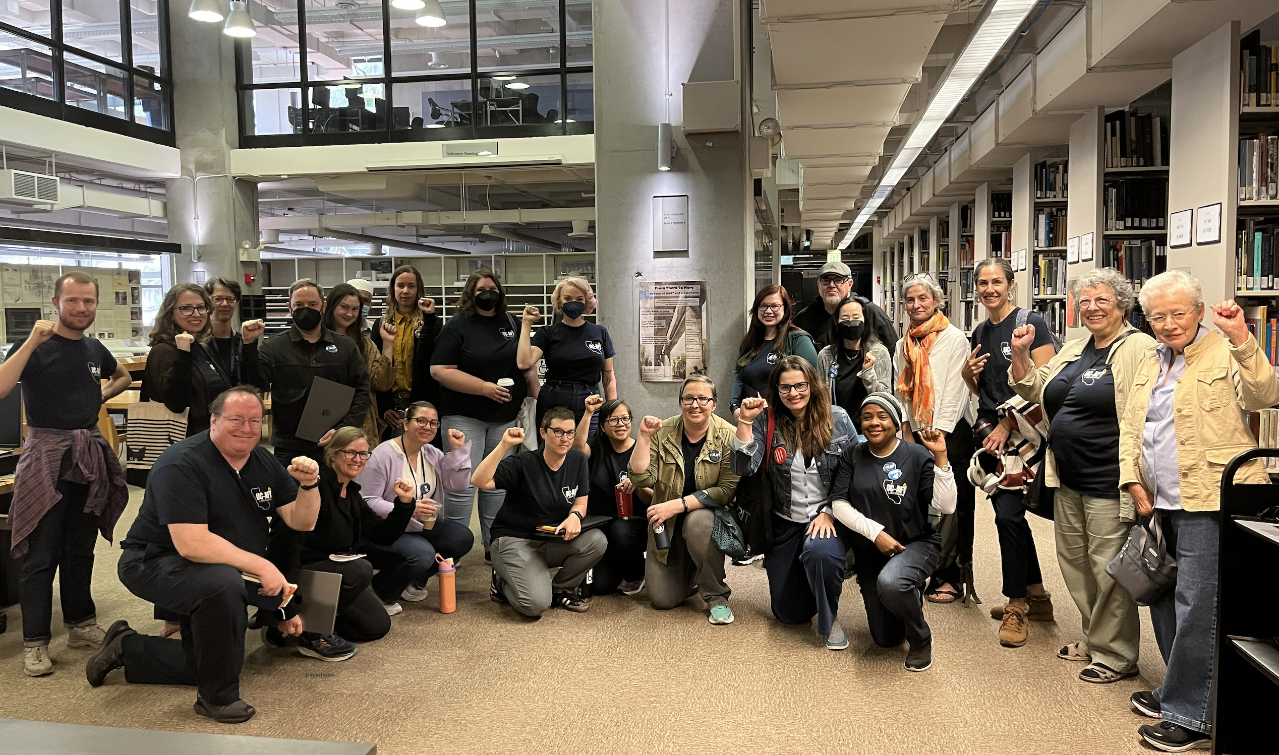 A group of approximately 25 people, of diverse ages and backgrounds, are gathered in a library setting. They are standing and kneeling, facing the camera, with most raising their fists in a gesture of solidarity.